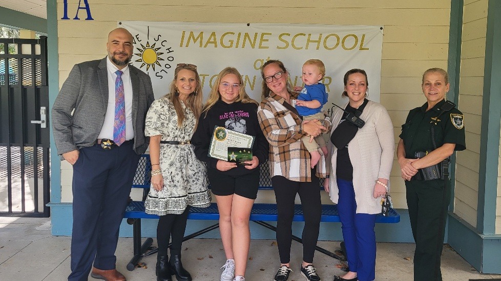 A group of seven people, including two girls (one holding a certificate), two women (one holding a baby), a man in a suit, and a uniformed police officer, stand smiling in front of an "Imagine School" banner