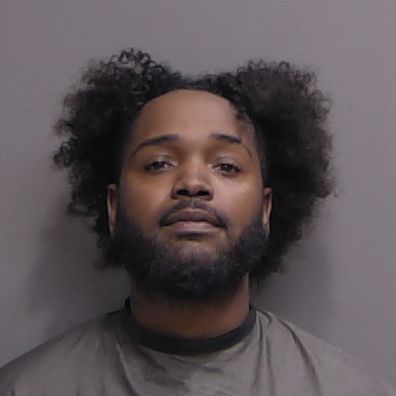 A man with a beard and mustache, wearing a gray garment, stands against a plain background His curly hair is parted outward, and he looks directly at the camera with a neutral expression