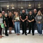 A group of adults, including uniformed officers and civilians, pose smiling indoors in front of a trophy case One woman in the center holds a certificate, and two people hold green and white pom poms