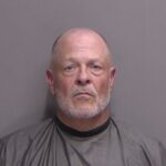 A middle aged man with a bald head, gray beard, and mustache poses against a plain gray background, wearing a dark, draped garment similar to a barber or mugshot cape