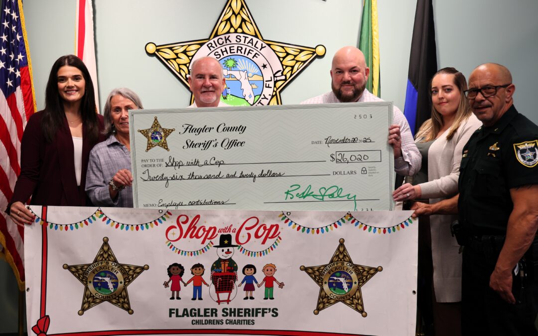 Six people stand together holding an oversized check for $, from the Flagler County Sheriff’s Office for "Shop with a Cop " They are behind a banner with sheriff stars and children's drawings Flags are visible on both sides