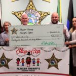 Six people stand together holding an oversized check for $, from the Flagler County Sheriff’s Office for "Shop with a Cop " They are behind a banner with sheriff stars and children's drawings Flags are visible on both sides