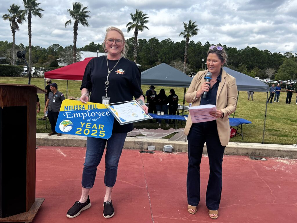 A smiling woman holding an "Employee of the Year " sign and a certificate stands next to another woman speaking into a microphone outdoors, with tents, people, and palm trees in the background A smiling woman holding an "Employee of the Year " sign and a certificate stands next to another woman speaking into a microphone outdoors, with tents, people, and palm trees in the background