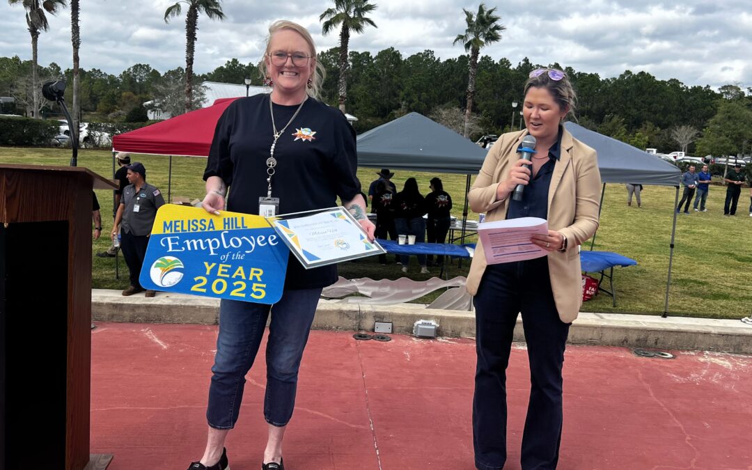 A smiling woman holding an "Employee of the Year " sign and a certificate stands next to another woman speaking into a microphone outdoors, with tents, people, and palm trees in the background