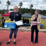 A smiling woman holding an "Employee of the Year " sign and a certificate stands next to another woman speaking into a microphone outdoors, with tents, people, and palm trees in the background