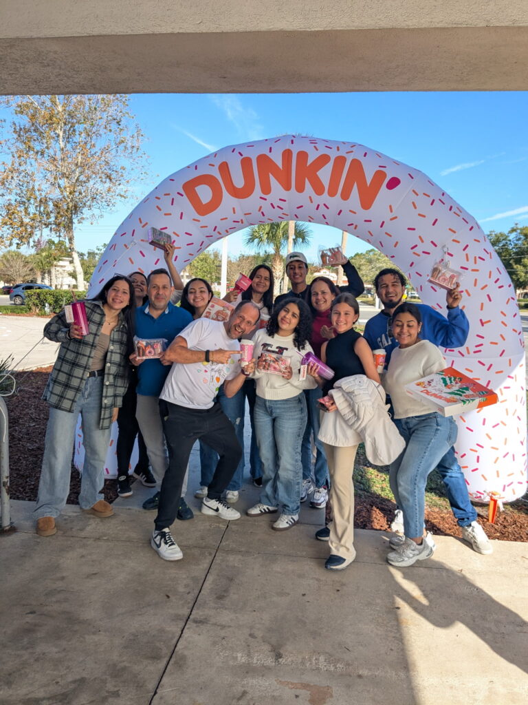 A group of people smiling and posing under a large inflatable Dunkin’ arch outdoors, holding Dunkin’ drinks and boxes of donuts Trees and a clear sky are visible in the background