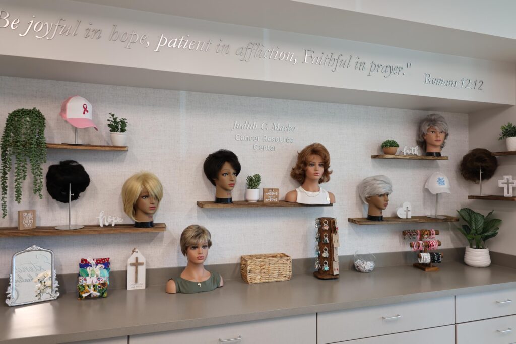 A display of wigs on mannequin heads is arranged on shelves in a resource center, along with hats, inspirational plaques, and support bracelets A Bible verse is inscribed on the wall above the shelves A display of wigs on mannequin heads is arranged on shelves in a resource center, along with hats, inspirational plaques, and support bracelets A Bible verse is inscribed on the wall above the shelves