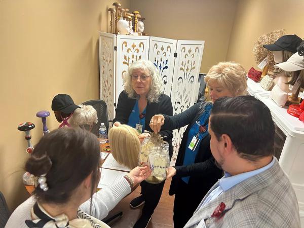 Four people gather in a small room with wigs, mannequins, and hair accessories Two women demonstrate a wig or hairpiece, while others observe and participate The setting appears professional and collaborative