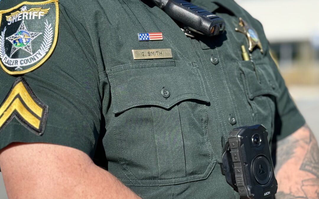 A close up of a sheriff's deputy in uniform wearing a body camera and radio The nameplate reads "G Smith" and the badge indicates Blazer County An American flag patch is visible above the nameplate