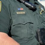 A close up of a sheriff's deputy in uniform wearing a body camera and radio The nameplate reads "G Smith" and the badge indicates Blazer County An American flag patch is visible above the nameplate