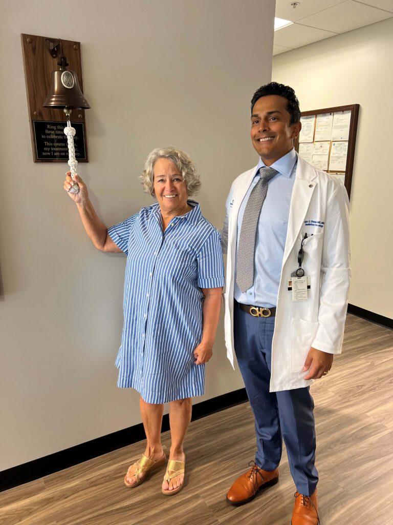 A smiling woman in a blue striped dress rings a bell on the wall, standing next to a doctor in a white coat They are both inside a medical office with certificates visible on the wall A smiling woman in a blue striped dress rings a bell on the wall, standing next to a doctor in a white coat They are both inside a medical office with certificates visible on the wall