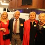 Four adults, three women and one man, stand together smiling at an indoor event They are dressed in formal attire with name tags, and a bar with drinks is visible in the background