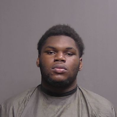 A young man with short, dark hair and a trimmed beard faces forward with a neutral expression, wearing a dark, cape like drape against a plain gray background