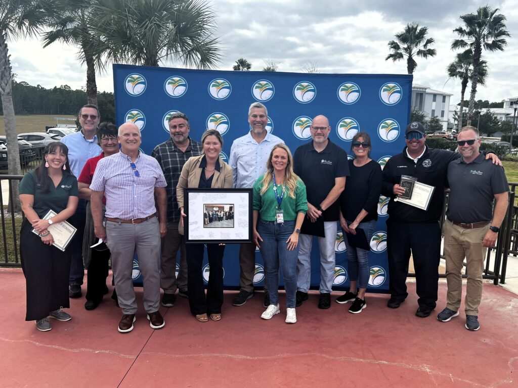 A group of twelve people stands outdoors in front of a blue backdrop with logos Some hold certificates or a framed photo Palm trees and clouds are in the background The group is smiling and posing for the picture A group of twelve people stands outdoors in front of a blue backdrop with logos Some hold certificates or a framed photo Palm trees and clouds are in the background The group is smiling and posing for the picture