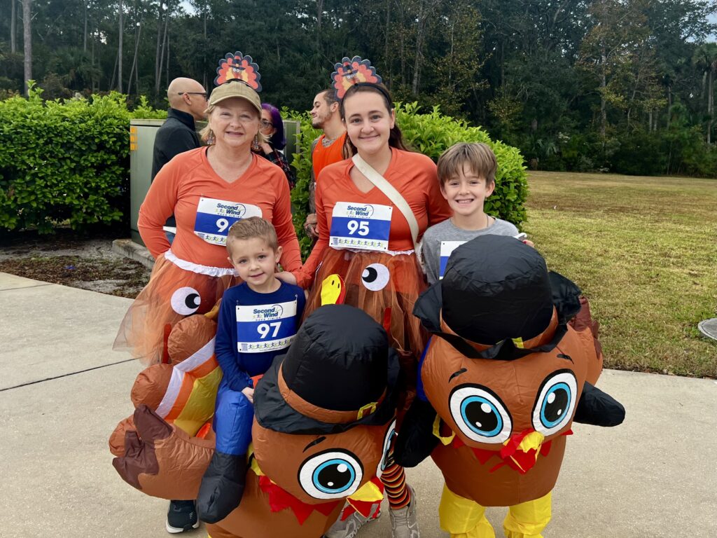 Four people, including two children and two adults, wear turkey costumes and race bibs while smiling outdoors at a fun run event Bushes and trees are visible in the background