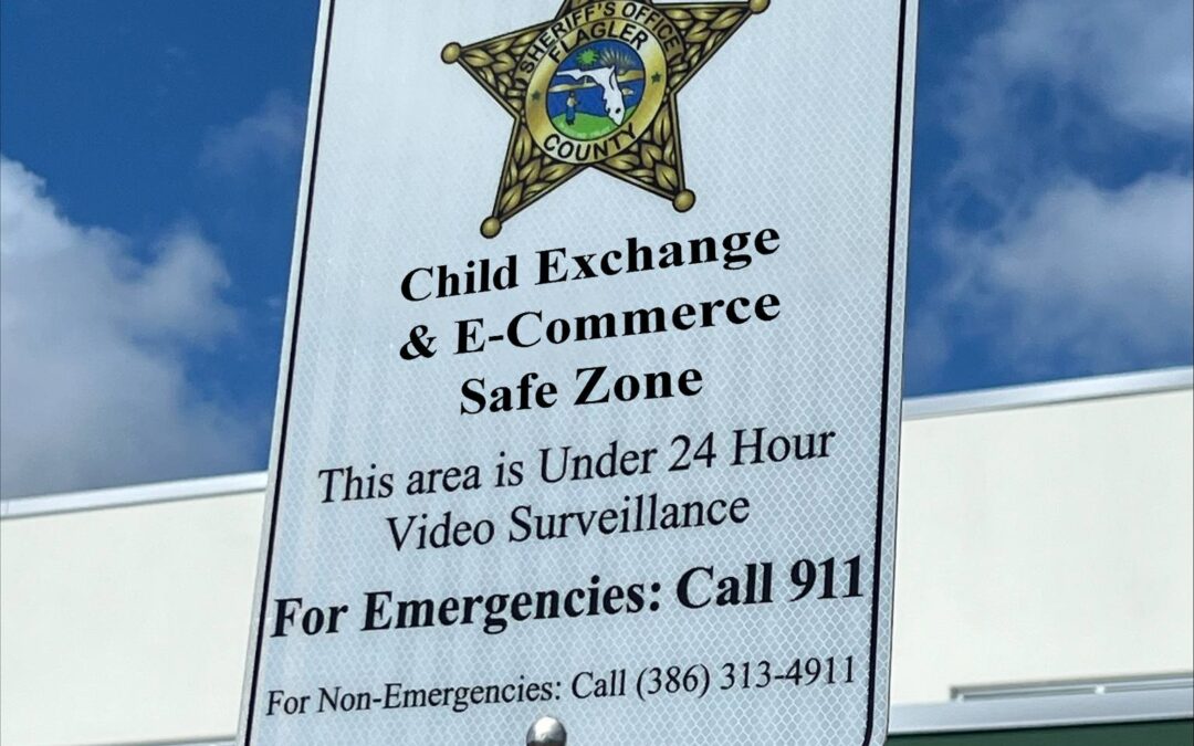 A sign with a sheriff's badge reads: "Child Exchange & E Commerce Safe Zone This area is under hour video surveillance For Emergencies: Call For Non Emergencies: Call ()