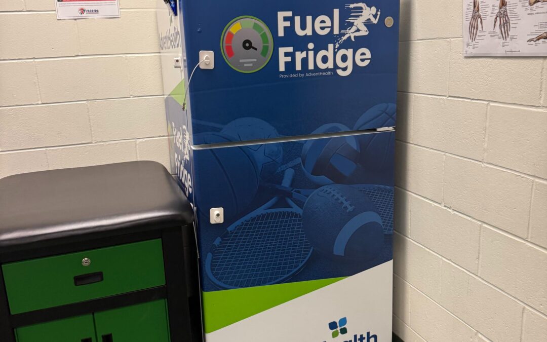 A blue and white "Fuel Fridge" by AdventHealth stands in a room with sports posters on the wall The fridge has images of various sports balls and is next to a green and black cabinet