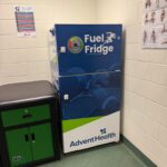 A blue and white "Fuel Fridge" by AdventHealth stands in a room with sports posters on the wall The fridge has images of various sports balls and is next to a green and black cabinet