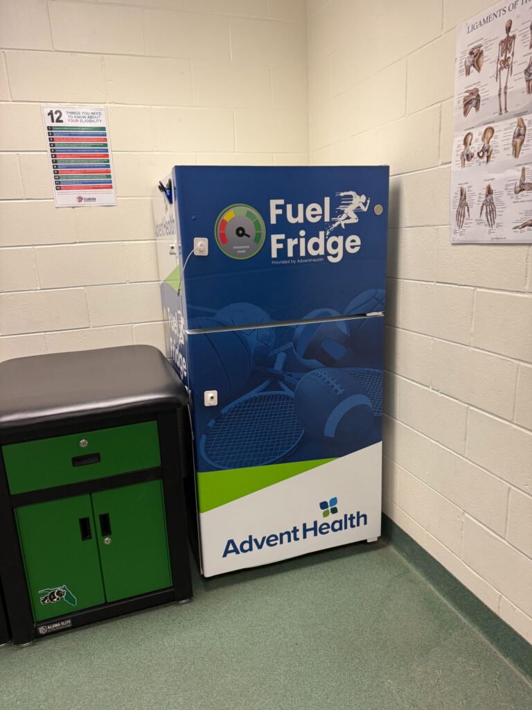 A blue and white "Fuel Fridge" by AdventHealth stands in a room with sports posters on the wall The fridge has images of various sports balls and is next to a green and black cabinet A blue and white "Fuel Fridge" by AdventHealth stands in a room with sports posters on the wall The fridge has images of various sports balls and is next to a green and black cabinet