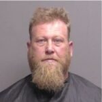 A middle aged man with a light brown beard and short hair is posed against a neutral background, wearing a dark drape typically used for mugshots