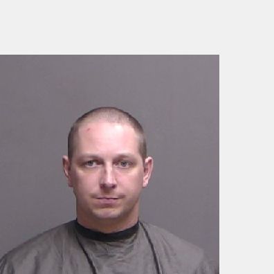A man with short hair wearing a gray garment poses in front of a plain gray background for a mugshot style photo