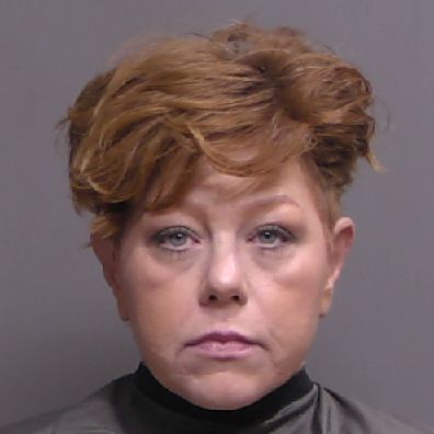 A woman with short, tousled red hair and light skin looks directly at the camera with a neutral expression, wearing a dark cape or drape around her shoulders against a plain background