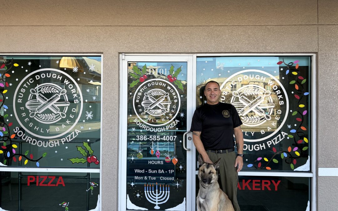A man in uniform stands beside a dog in front of a storefront with "Rustic Dough Works Sourdough Pizza" logos and holiday decorations on the doors and windows A menorah and business hours are visible on the glass