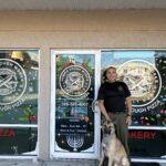 A man in uniform stands beside a dog in front of a storefront with "Rustic Dough Works Sourdough Pizza" logos and holiday decorations on the doors and windows A menorah and business hours are visible on the glass