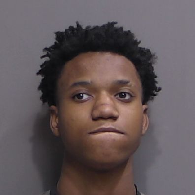 A young person with short, curly hair looks slightly upward with a neutral expression while biting their lower lip, set against a plain, grey background