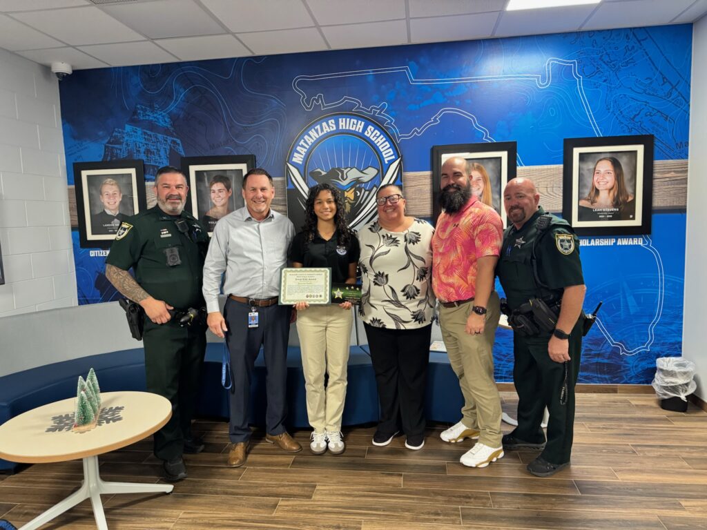 Six adults stand in a school lobby in front of a blue mural and framed photos A young woman in the center holds a certificate, smiling, surrounded by school staff and uniformed officers