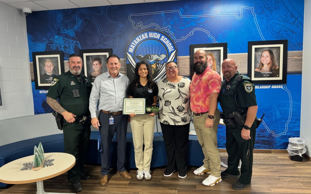 Six adults stand in a school lobby in front of a blue mural and framed photos A young woman in the center holds a certificate, smiling, surrounded by school staff and uniformed officers