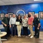Six adults stand in a school lobby in front of a blue mural and framed photos A young woman in the center holds a certificate, smiling, surrounded by school staff and uniformed officers