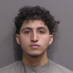 A young man with curly dark hair and slight facial hair stares directly at the camera with a neutral expression, wearing a gray drape against a plain gray background