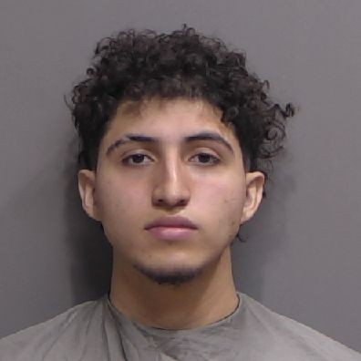 A young man with curly dark hair and slight facial hair stares directly at the camera with a neutral expression, wearing a gray drape against a plain gray background
