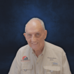 An older man with short gray hair smiles at the camera against a dark blue background He is wearing a beige shirt with Flagler patches and embroidery on it