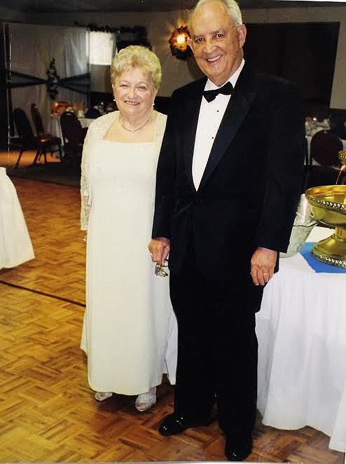 An older woman in a light dress and an older man in a black tuxedo stand together, smiling, on a parquet floor at the elegant Flagler banquet hall Tables with white tablecloths are visible in the background An older woman in a light dress and an older man in a black tuxedo stand together, smiling, on a parquet floor at the elegant Flagler banquet hall Tables with white tablecloths are visible in the background