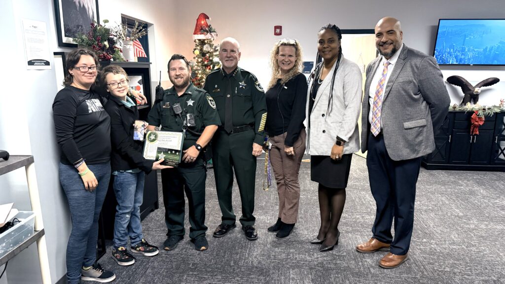 A group of seven people, including two uniformed sheriffs, pose indoors One sheriff holds a certificate, standing next to a woman and child The group smiles, and festive decorations are visible in the background
