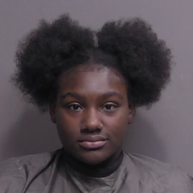 A young person with dark skin and natural hair styled in two large puffs looks straight at the camera against a plain background, wearing a dark, draped garment