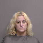 A woman with light blonde, wavy hair and a neutral expression poses in front of a plain gray background, wearing a dark cape typically used in mugshot photos