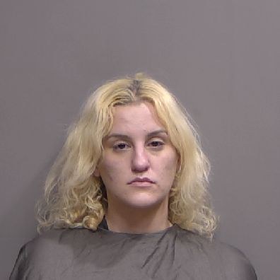 A woman with light blonde, wavy hair and a neutral expression poses in front of a plain gray background, wearing a dark cape typically used in mugshot photos A woman with light blonde, wavy hair and a neutral expression poses in front of a plain gray background, wearing a dark cape typically used in mugshot photos