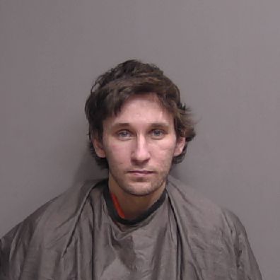 A young man with tousled brown hair and light facial hair is wearing a grayish poncho like garment, standing against a plain gray background, looking directly at the camera with a neutral expression