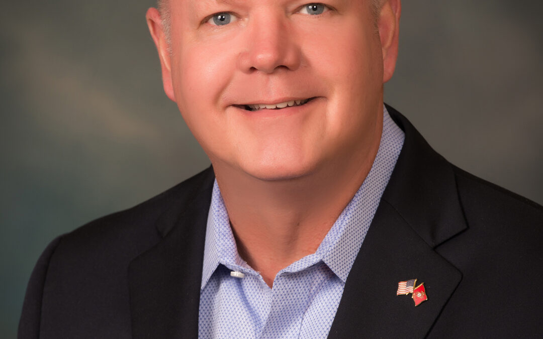 A middle aged man with short, light hair smiles at the camera He wears a dark blazer, light blue shirt, and a small American flag pin with another red pin on his lapel, representing Flagler County The background is softly blurred