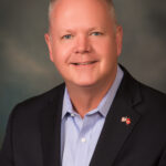 A middle aged man with short, light hair smiles at the camera He wears a dark blazer, light blue shirt, and a small American flag pin with another red pin on his lapel, representing Flagler County The background is softly blurred