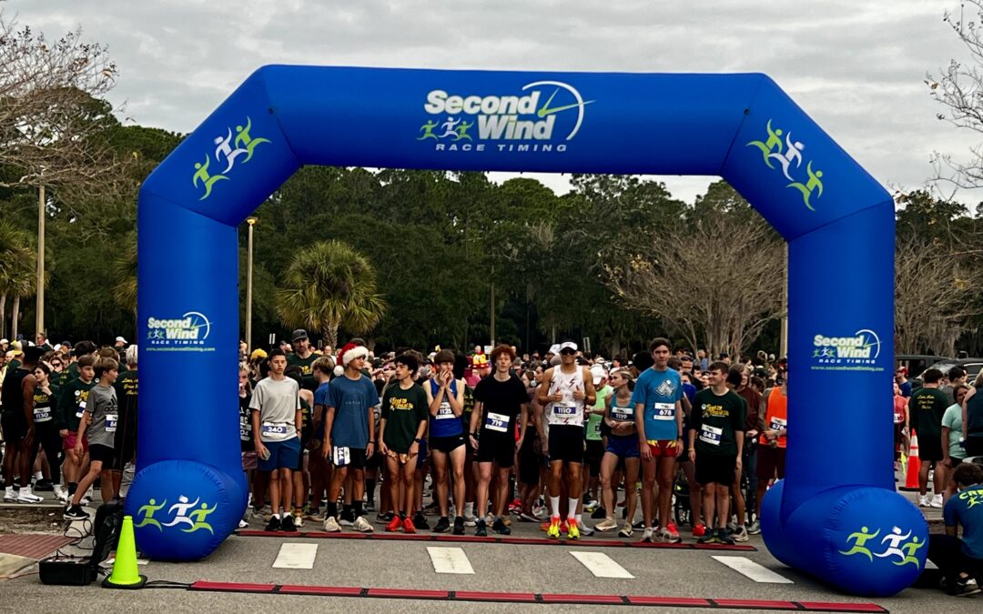 A group of runners stands at the starting line beneath a large blue race arch labeled "Second Wind Race Timing," preparing to begin a race on a cloudy day with trees in the background
