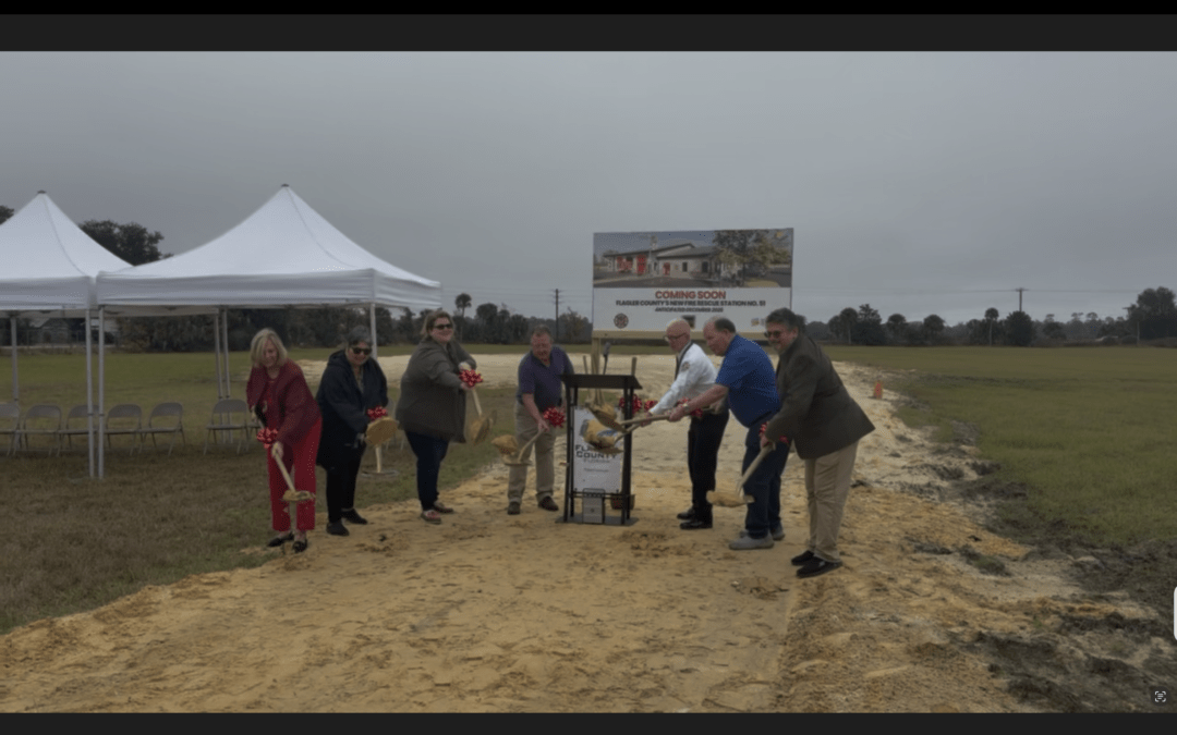 Seven people participate in a groundbreaking ceremony on a construction site, holding shovels with red bows Two white tents and a sign with building plans are in the background, with a grassy field and cloudy sky
