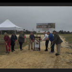 Seven people participate in a groundbreaking ceremony on a construction site, holding shovels with red bows Two white tents and a sign with building plans are in the background, with a grassy field and cloudy sky