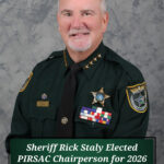 A smiling sheriff in uniform with badges, pins, and medals poses against a gray backdrop Text on the image reads: "Sheriff Rick Staly Elected PIRSAC Chairperson for