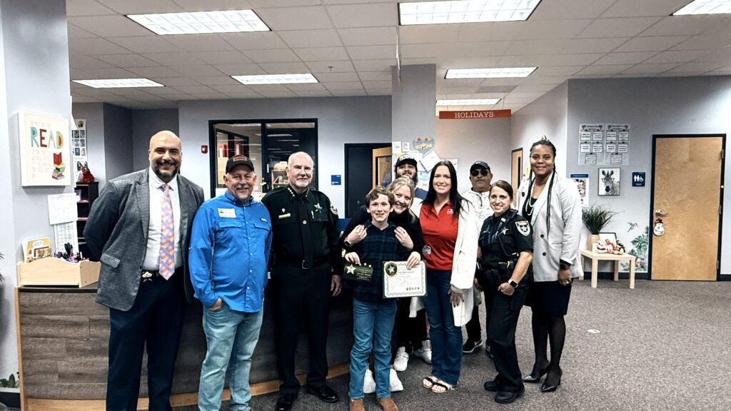 A group of people, including law enforcement officers, school staff, and children, pose together indoors One child holds a certificate, and everyone is smiling The setting appears to be a school office area