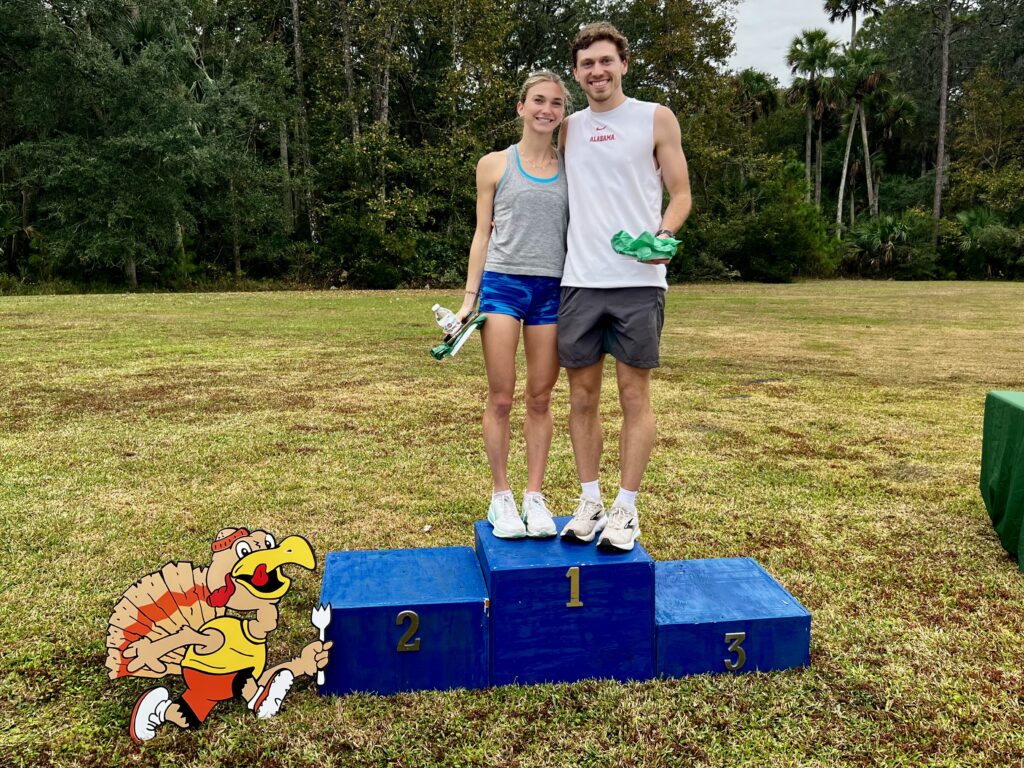 A smiling young woman and man stand on a blue podium’s top spot outdoors, holding green prizes The woman is on spot , the man on spot A cartoon turkey wearing running shoes is on the grass next to the podium
