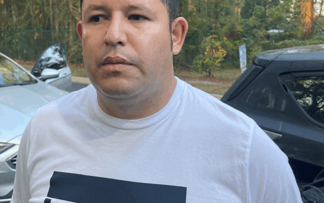 A man with short dark hair wearing a white Levi's t shirt stands outdoors in front of parked cars and trees with autumn foliage